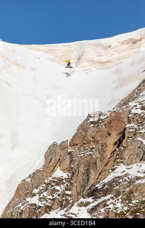 A snowboarder in a vibrant yellow jacket jumps off a steep snow-covered cliff, showcasing extreme freeride skills in pristine backcountry powder again Stock Photo