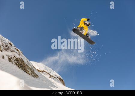 A snowboarder in a yellow jacket performs a thrilling jump in the backcountry, showcasing a breathtaking freeride experience against a clear blue sky Stock Photo