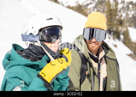 Two snowboarders equipped with gear enjoying a sunny day in the snowy backcountry. They are dressed for cold conditions, wearing helmets and goggles, Stock Photo