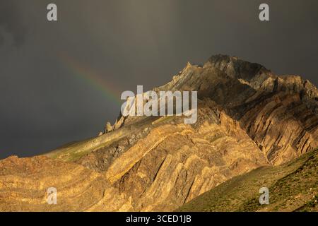 A breathtaking view of a rugged mountain in Spiti Valley, India, adorned with a delicate rainbow against a dramatic sky. The terrain showcases the reg Stock Photo
