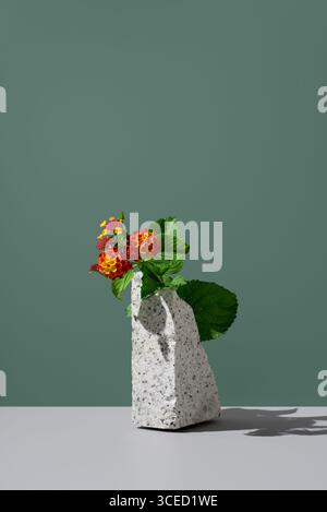 Red and yellow Lantana camara flowers in a stone-textured vase on a solid gray surface against a muted green background. The vibrant blooms are from t Stock Photo