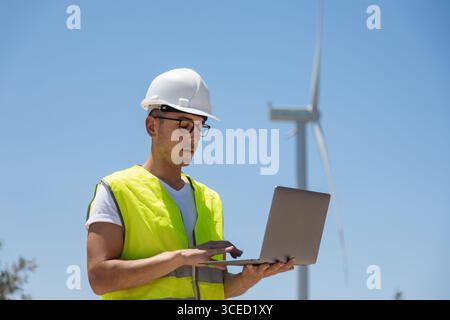An engineer wearing a hard hat and safety vest stands outdoors, using a laptop to inspect wind turbines. The bright blue sky and wind turbine in the b Stock Photo