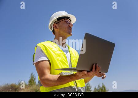 An engineer in a safety vest and helmet inspects wind turbines using a laptop in an open field, ensuring functionality and efficiency of renewable ene Stock Photo