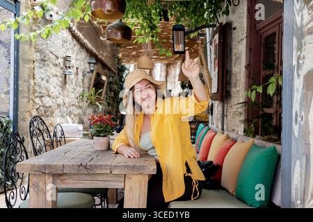 A young, cheerful woman in a yellow shirt sits at a cafe table, wearing a straw hat. She is smiling brightly and gesturing as she seems ready to order Stock Photo