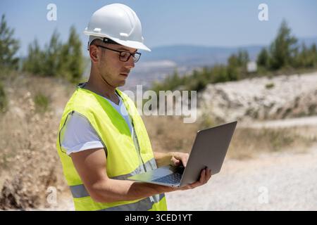 An engineer wearing a safety helmet and vest examines data on a laptop while inspecting wind turbines in a rural area. The professional is focused on Stock Photo