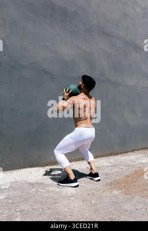 A fit man performs a wall exercise with a medicine ball, wearing athletic gear. His strong, muscular build is highlighted as he maintains balance and Stock Photo