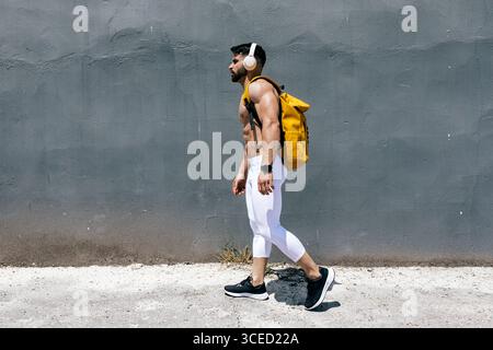 Fit man walking along a concrete path, wearing white leggings and black sneakers. He carries a yellow backpack and wears headphones, embodying a sport Stock Photo
