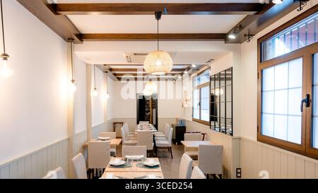 A modern restaurant interior with elegant decor. Wooden beams complement white walls, while soft pendant lights illuminate neatly set dining tables, c Stock Photo