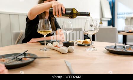 A woman pours white wine into a glass at a restaurant dinner setting. The table is elegantly set with empty plates and glasses, creating a cozy dining Stock Photo