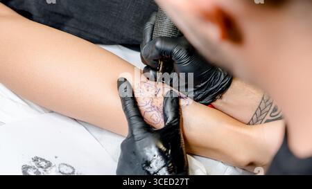 A tattoo artist skillfully applying an intricate design on a client’s leg using a precision needle, demonstrating expertise in the art of tattooing in Stock Photo