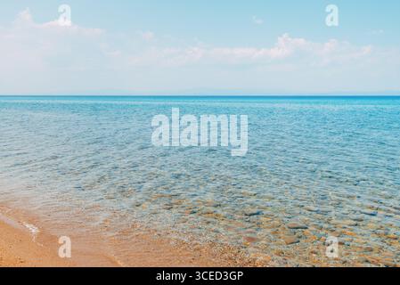 Clear turquoise sea with smooth stones visible beneath the shallow water. Calm shoreline under a bright sky, distant mountains faintly seen on the hor Stock Photo