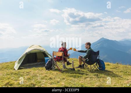 Couple relaxing in camping chairs beside their tent, enjoying a scenic mountain view and peaceful atmosphere under the blue sky. Stock Photo