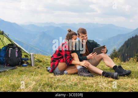 Modern young couple camping in the mountains, blending tech and nature, working remotely on a tablet while cooking outdoors and enjoying the views. Stock Photo