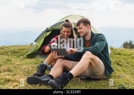 Modern young couple camping, enjoying time outside their tent, laughing, sharing moments, and streaming on a tablet in a stunning natural setting. Stock Photo