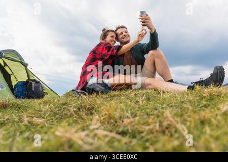 Cheerful couple having fun by their tent on a mountain camping spot, taking memorable selfie photos with a smartphone. Stock Photo