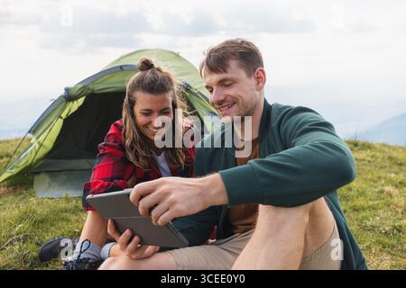 Smiling couple sitting outside their tent during a camping trip, watching a tablet screen and relaxing together in the great outdoors. Stock Photo