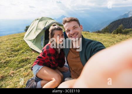 Couple enjoying a chill camping moment, taking selfies outside their tent with stunning mountain scenery in the backdrop. Stock Photo