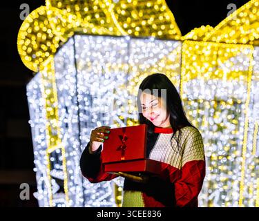 Woman smiling while opening red gift box under holiday lights. Christmas. Celebration. Stock Photo