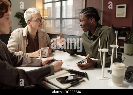 Group of three young ethnically diverse specialists sitting at office desk, discussing renewable energy solutions Stock Photo