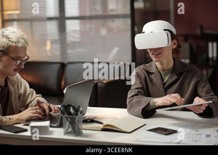 Young female manager using digital tablet and VR headset while her male colleague typing on laptop in modern office Stock Photo
