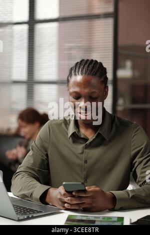 Vertical medium portrait of cheerful young Gen Z Black man sitting at office desk using smartphone Stock Photo