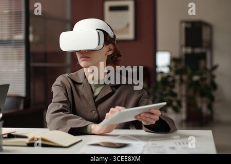 Gen Z Caucasian female specialist using VR headset and digital tablet at workplace in modern office Stock Photo