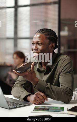 Medium shot of cheerful young Black man recording voice message via smartphone, while working in modern office Stock Photo