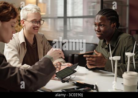 Group of three young ethnically diverse engineers sitting at office desk, discussing renewable energy solutions Stock Photo