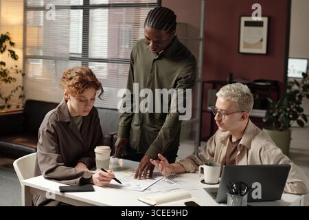 Group of three ambitious young coworkers discussing ideas while working on new project in modern office Stock Photo