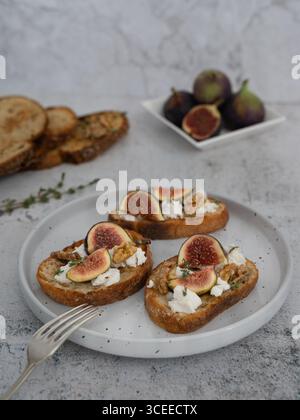 Front view vertical close up of three fig and goat cheese toasts on white plate with gray background, fork, figs on separate plate, toasts in backgrou Stock Photo
