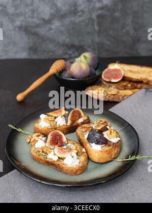 Top view vertical of three fig and goat cheese toasts on green plate with gray kitchen towel, honey dipper, figs, copy space, dark background Stock Photo