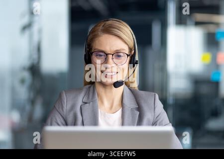 Smiling professional woman with headset using laptop during online meeting in modern office, symbolizing communication, support, teamwork, and corporate success. Stock Photo