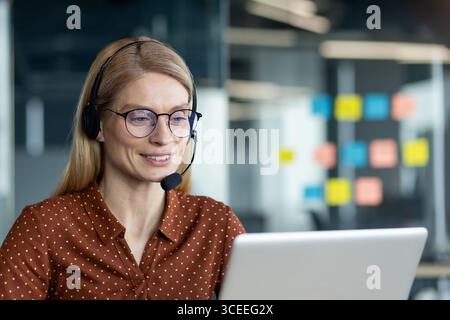 A confident businesswoman in glasses and headset reviewing tasks on a laptop, collaborating remotely. Stock Photo