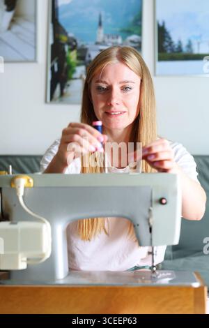 Young woman sewing clothes on retro sewing machine, smiling with joy. Stock Photo