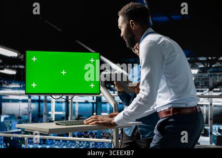 African american IT experts using chroma key device to analyze neural network systems. Data center admin uses green screen pc next to colleague, reviewing AI visualization in background. Stock Photo
