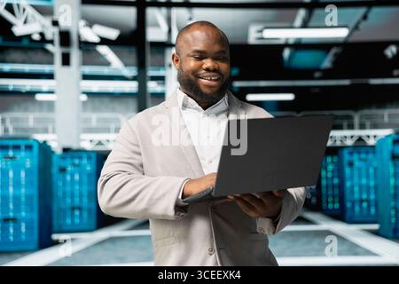 Happy programmer in server farm overseeing mainframes devices used for managing databases. Cheerful man in data center establishment doing checkup on rackmounts storing information Stock Photo