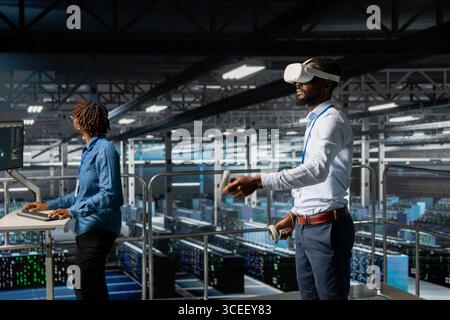 Black male IT expert uses VR glasses on industrial platform in server room, using high tech tools to manage machine learning for automation. Neural network systems in a data center. Stock Photo