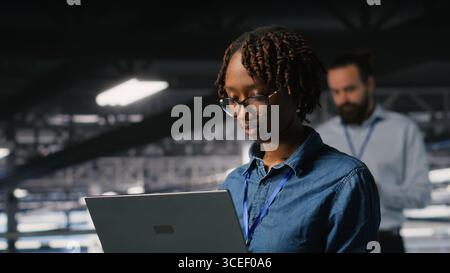 System administrators on data center industrial platform analyzing performance outputs on laptop. Server hub teamworking colleagues finetuning server rig configurations using notebook Stock Photo