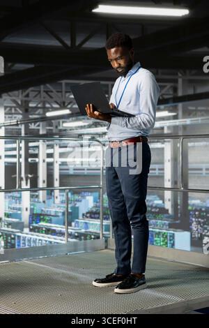 Server room black engineer monitoring artificial intelligence model with laptop, overseeing system performance infrastructure from industrial platform. Male technician reviews neural network. Stock Photo