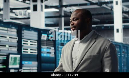 Smiling employee walking in high tech data center with machines powering networks, running companies databases. Admin overseeing server farm with rigs modules processing large datasets, camera B Stock Photo