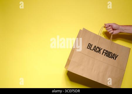 Black Friday brown paper shopping bag being held by a male hand on a yellow background with copy space. Stock Photo