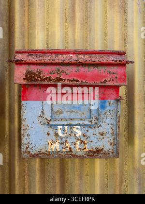 Close up of an old, rusty U.S. Mail box with chipped red and blue paint attached to a gold colored corrugated metal building. Stock Photo