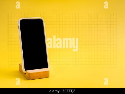 Smartphone with blank black screen on wooden stand against yellow background with grid, ideal for app presentation, mobile technology advertising, UI Stock Photo
