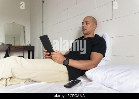 Middle aged man in black t-shirt relaxing on bed while using digital tablet computer in a minimalistic white apartment. Casual lifestyle scene in a mo Stock Photo