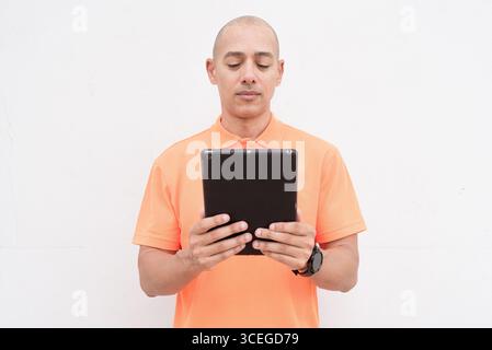 Studio portrait of handsome middle aged bald Hispanic man in orange polo shirt against plain white wall background using digital tablet computer. Casu Stock Photo