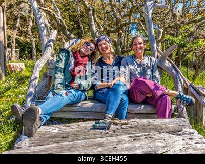 Three women friends sitting and smiling on a hand-built driftwood seat on Hornby Island, British Columbia, Canada. Stock Photo