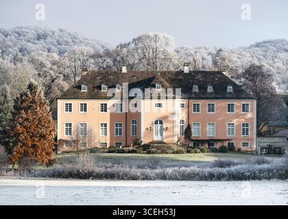 Charming historic mansion with a peach facade stands quietly in a snowy winter landscape. Frost-covered trees and bushes surround the house. Stock Photo