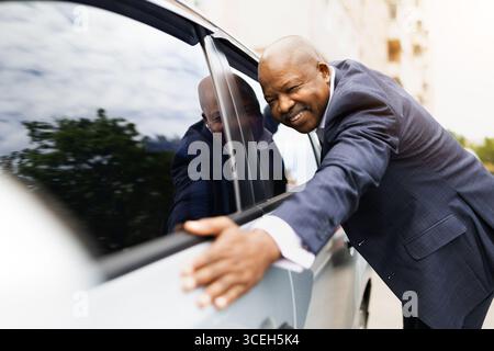 Smiling black businessman standing outside car leaning on door, embracing new vehicle Stock Photo
