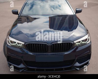 Close-up front view of shiny, modern dark blue sedan. The photo highlights grille, headlights, with reflections of the urban buildings on car hood. Stock Photo