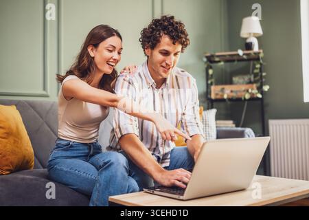 Adult couple is sitting at sofa at home and using laptop. They are shopping online and searching for goods. Stock Photo